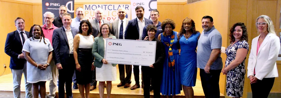 A group of people stand in front of signage for Montclair Public Library and PSE&G, and two people hold up a big check from PSE&G to the library.