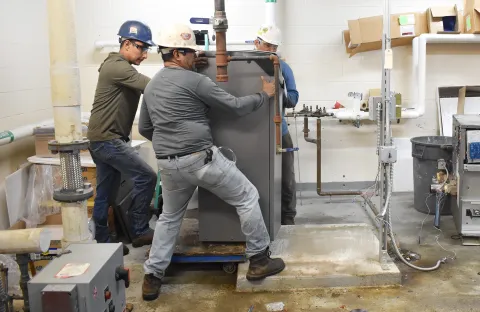 Technicians wearing hard hats use a wheeled platform to move a large mechanical unit inside an indoor utility room.