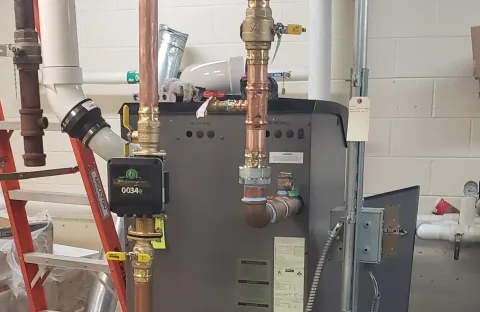 Indoor boiler connected to copper piping and valves in a utility room.